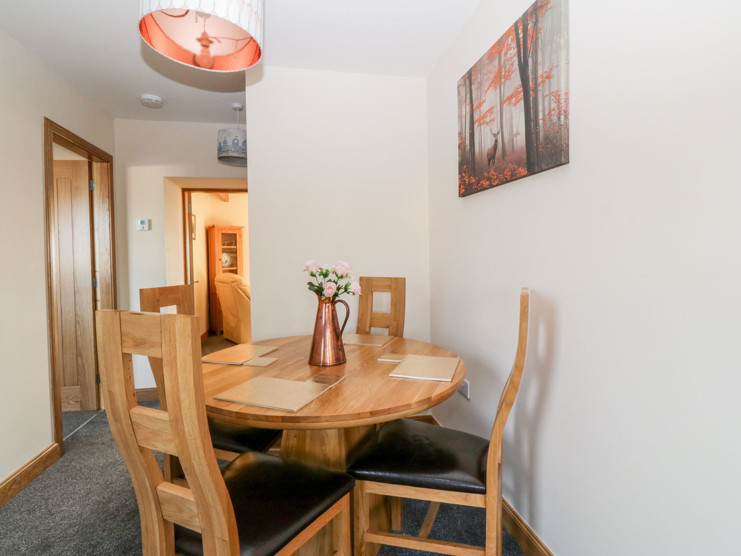A round wooden dining table with four chairs, a copper pitcher with flowers, and a painting of a forest scene on the wall.