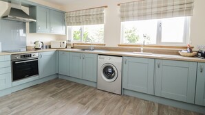 Una cocina moderna con armarios azul claro, horno empotrado, microondas y lavavajillas. Hay dos ventanas con cortinas a cuadros, y un lavabo con grifo.