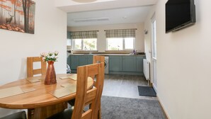 Un comedor con una mesa de madera, sillas y un jarrón de flores. Se observa una cocina al fondo, con una ventana y un televisor de pantalla plana montado en la pared.