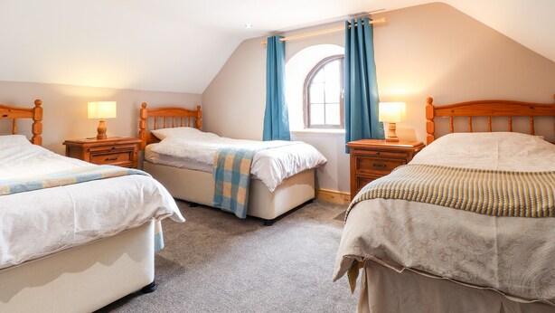A cozy attic bedroom with three wooden beds, a large window with blue curtains, and a sloped ceiling.