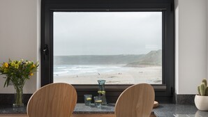 A modern kitchen with a large window overlooking a beach. There are two wooden chairs and a small table with a vase of flowers and two glasses on the counter.