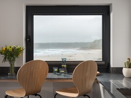 A modern kitchen with a large window overlooking a beach. There are two wooden chairs and a small table with a vase of flowers and two glasses on the counter.