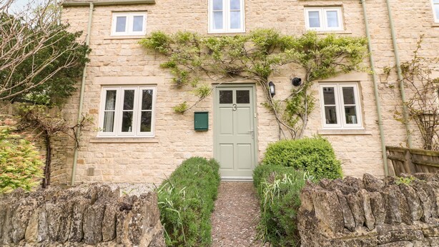 A stone house with a green door, two windows on the upper floor, and a small garden with a hedge and a tree.