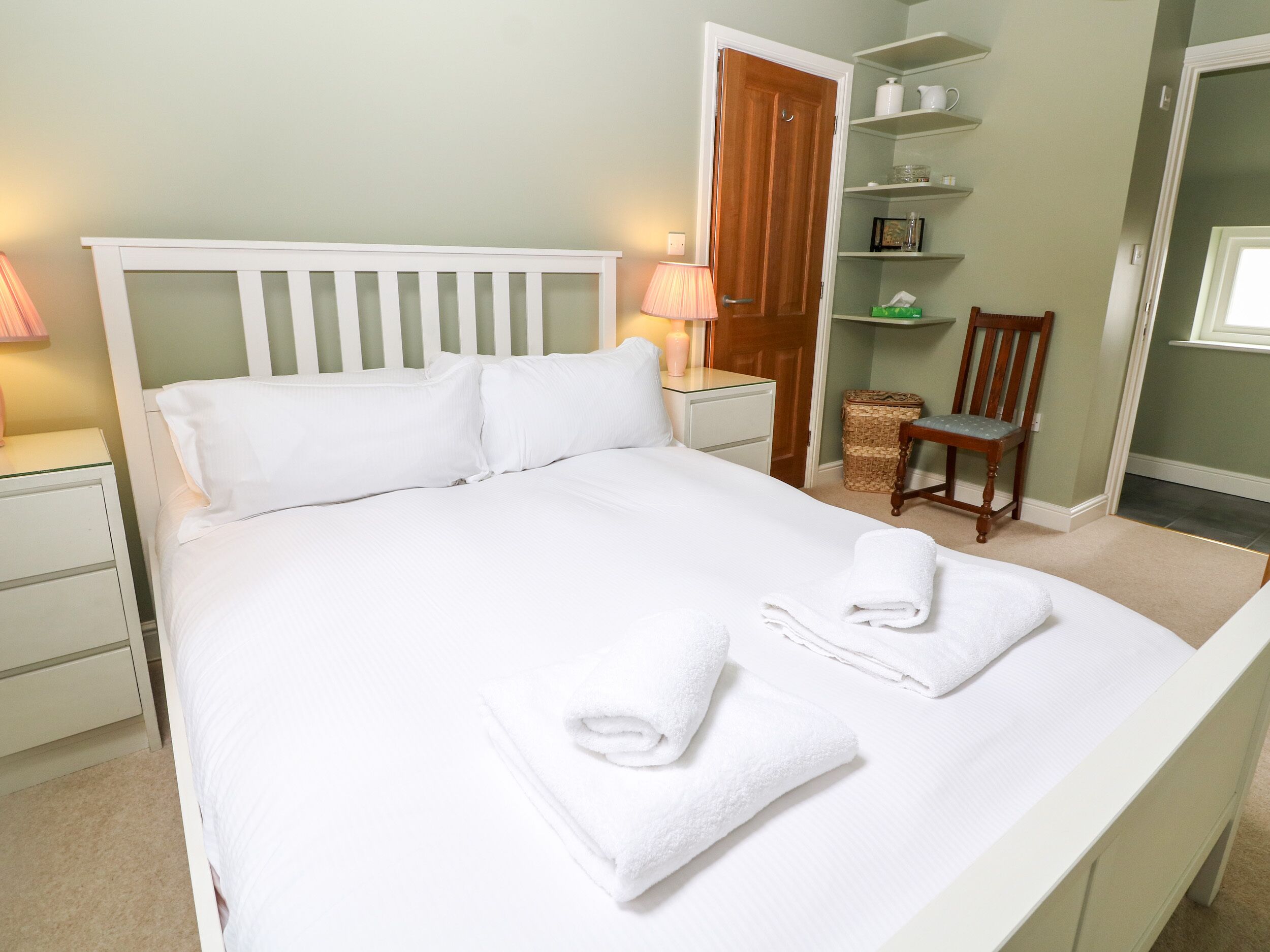 A neatly made bed with white linens and two folded towels, a bedside lamp, a wooden chair, and a small wicker basket.