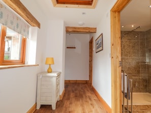 Um corredor com piso de madeira, um aparador branco com abajur, uma porta de madeira e um espaço de chuveiro com paredes revestidas de azulejos.