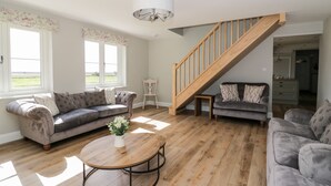 A spacious living room with a wooden staircase, two sofas, a coffee table, and a view of the outdoors through large windows. - West Farm (Great Yarmouth)