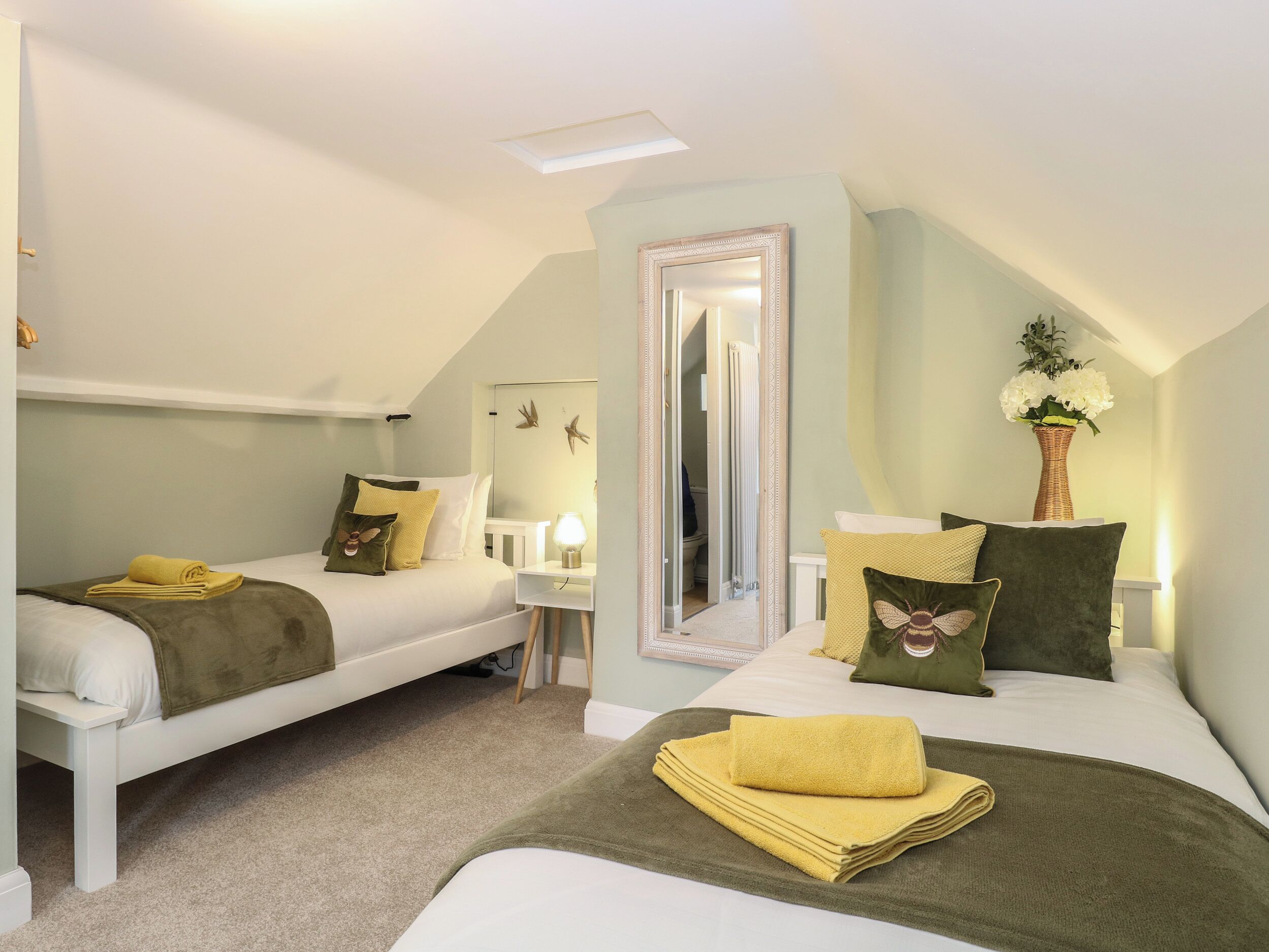 Une chambre d’hôtel avec deux lits, un miroir, une petite table avec une lampe et un vase avec des fleurs.