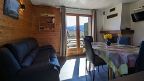Een gezellige kamer met houten inrichting, een bank, een eettafel met stoelen, een aan de muur bevestigde televisie en uitzicht op de omgeving door een glazen deur.