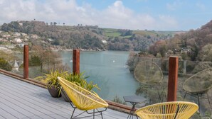 A terrace with wicker chairs and a table overlooking a lake with a village in the background. - High Banks (Dartmouth)