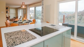 A modern kitchen with a central island, a dining area with chairs, and large windows offering a view of the countryside. - High Banks (Dartmouth)