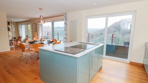 A modern kitchen with a central island, dining area with chairs, and large windows offering a view of the outdoors. - High Banks (Dartmouth)