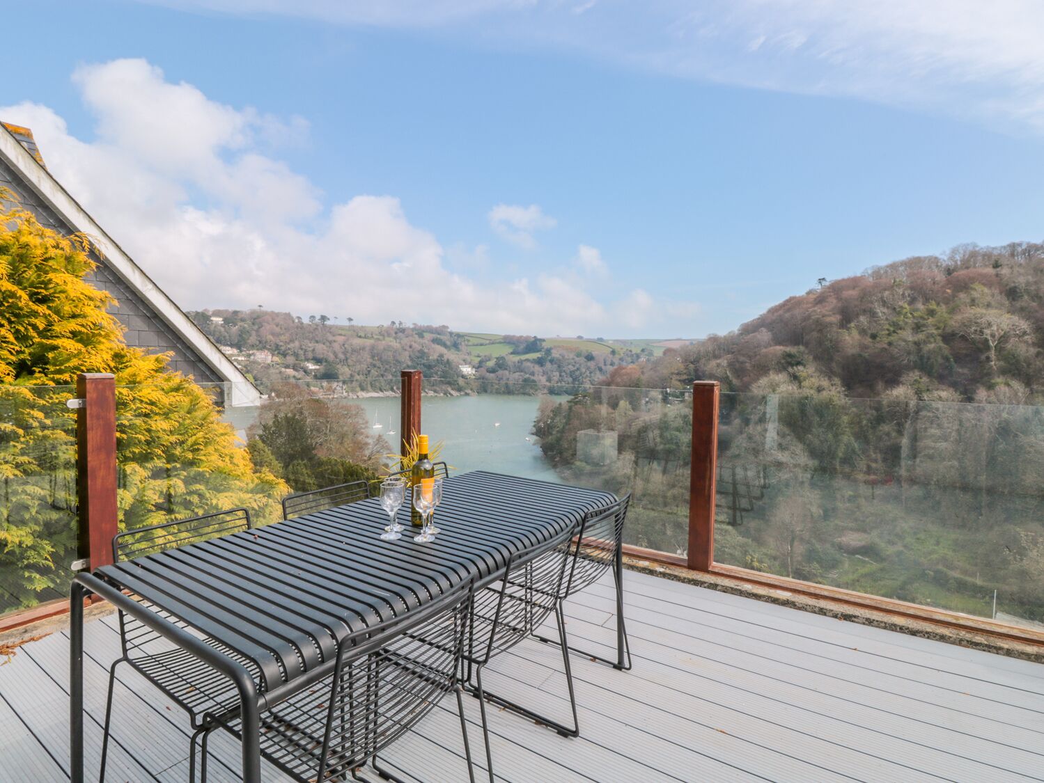 A table with a bottle and glasses on a deck with a view of a river and hills.
