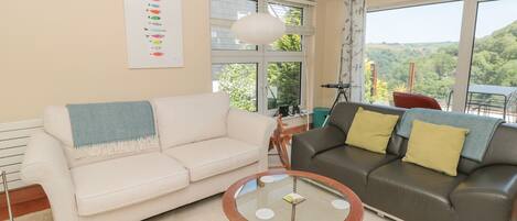 A living room with two sofas, a glass coffee table, and large windows offering a view of greenery.