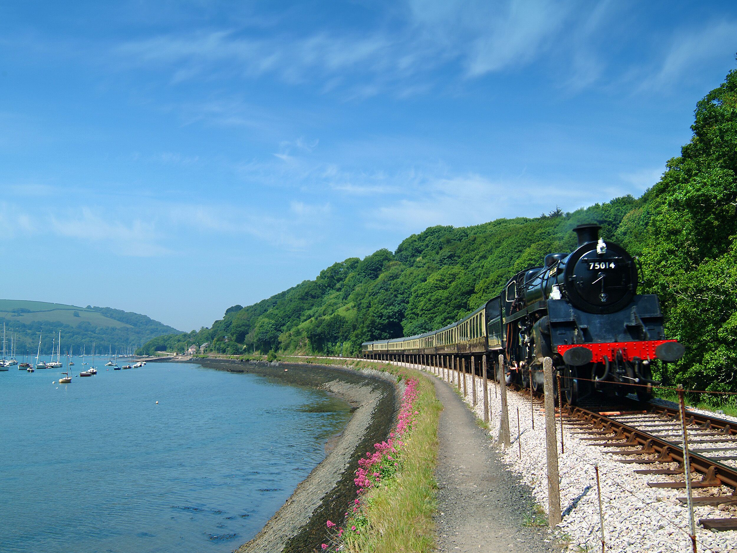 Un treno sui binari, accanto a un corso d'acqua con barche, un sentiero e vegetazione.