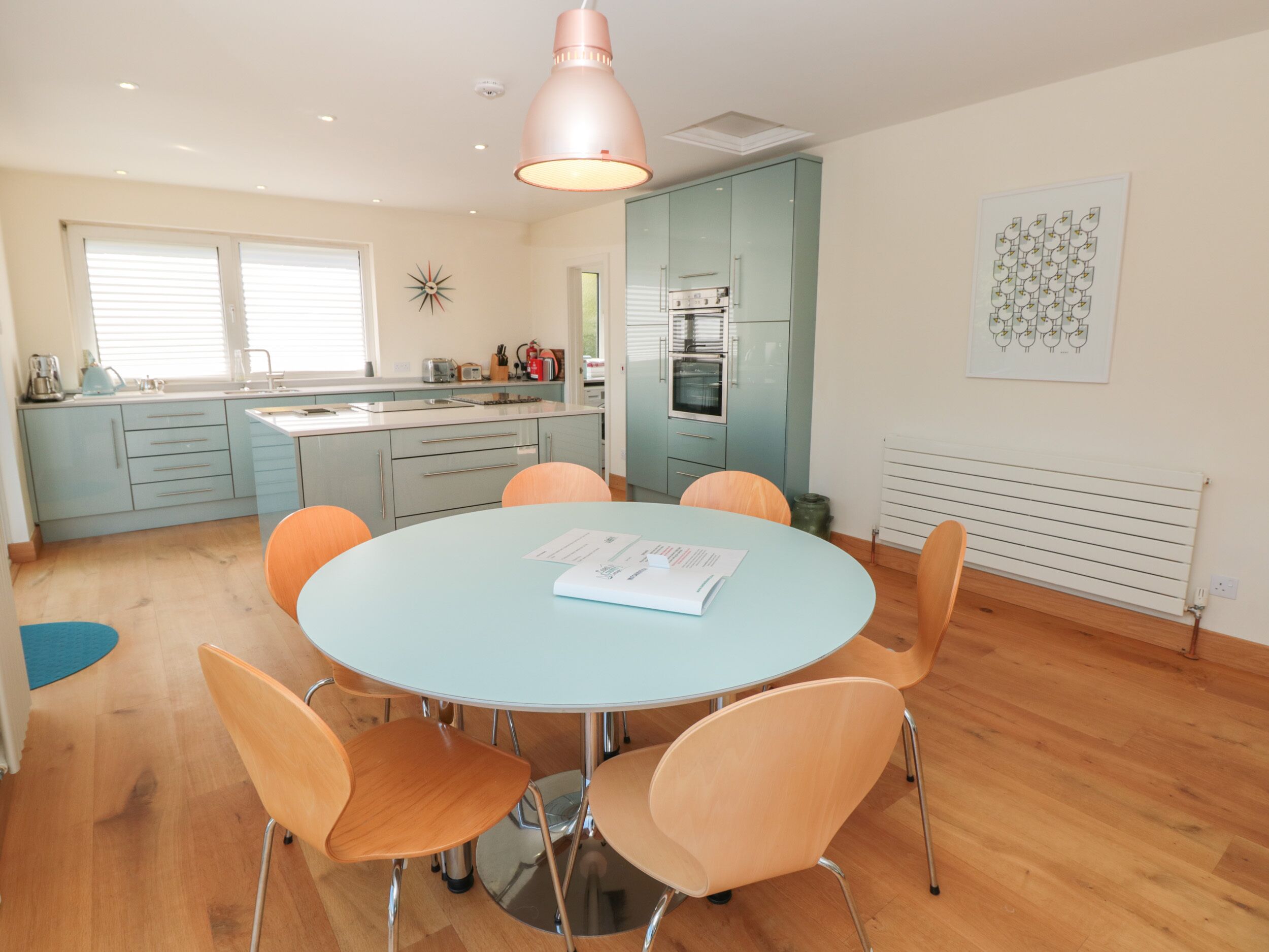 A modern kitchen with light blue cabinets, a round table with orange chairs, and a pendant light above.