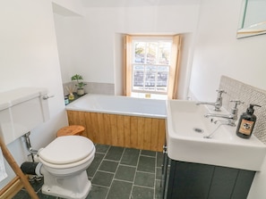Een badkamer met een wit toilet, een houten badkuip, een witte wastafel en een raam met uitzicht op gebouwen.