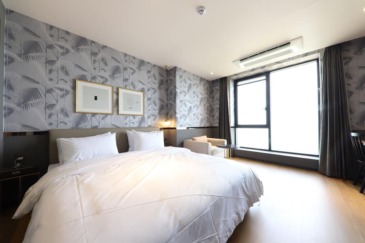 Una habitación de hotel moderna con una cama grande, una mesita de noche, un escritorio y un amplio ventanal con cortinas.