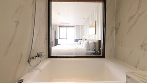 Une salle de bain moderne avec une grande baignoire, une douche et un miroir qui reflète une chambre à coucher avec un lit et une table de chevet.
