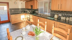 A kitchen with wooden cabinets, a black countertop, a sink, and a window with blinds. - Little Brook Cottage (Carmarthen)