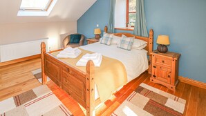 A bedroom with a sloped ceiling, a bed with a wooden headboard, two bedside tables with lamps, a chair, and a window with curtains. - Little Brook Cottage (Carmarthen)