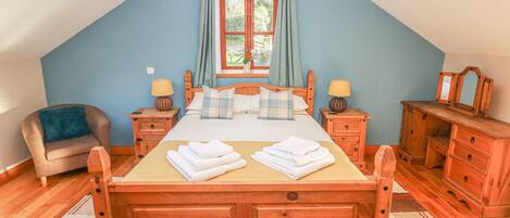 A bedroom with a sloped ceiling, a bed with a wooden headboard, two bedside tables with lamps, a chair, a dresser, and a window with curtains.