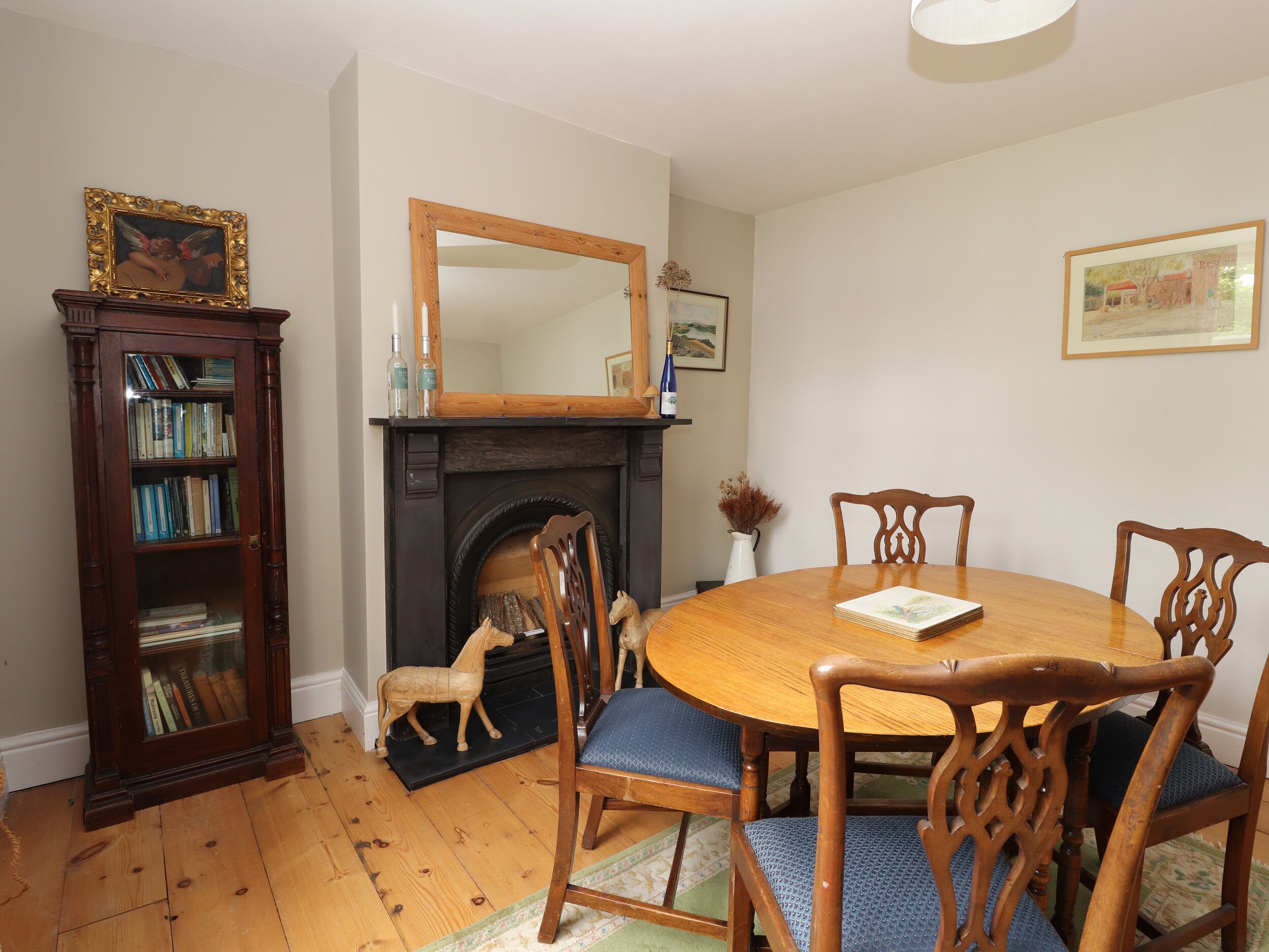 A dining room with a round wooden table, six chairs, a fireplace, a bookshelf, and a mirror on the wall.