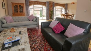 A living room with a grey sofa, a black sofa, and a coffee table with a book and remote control. There is a wooden cabinet and a dining area visible through a glass door. - Pipistrelle Cottage (Beverley)