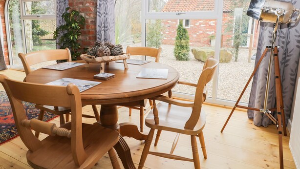 A dining area with a wooden table and chairs, a rustic brick wall, and large windows offering a view of the garden. - Pipistrelle Cottage (Beverley)