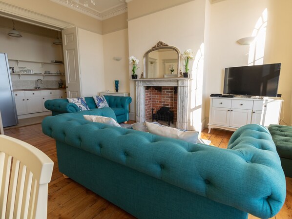 A living room with a teal sofa, a fireplace, a television, and a kitchen area visible through an open door. - Wessex View (Weymouth)