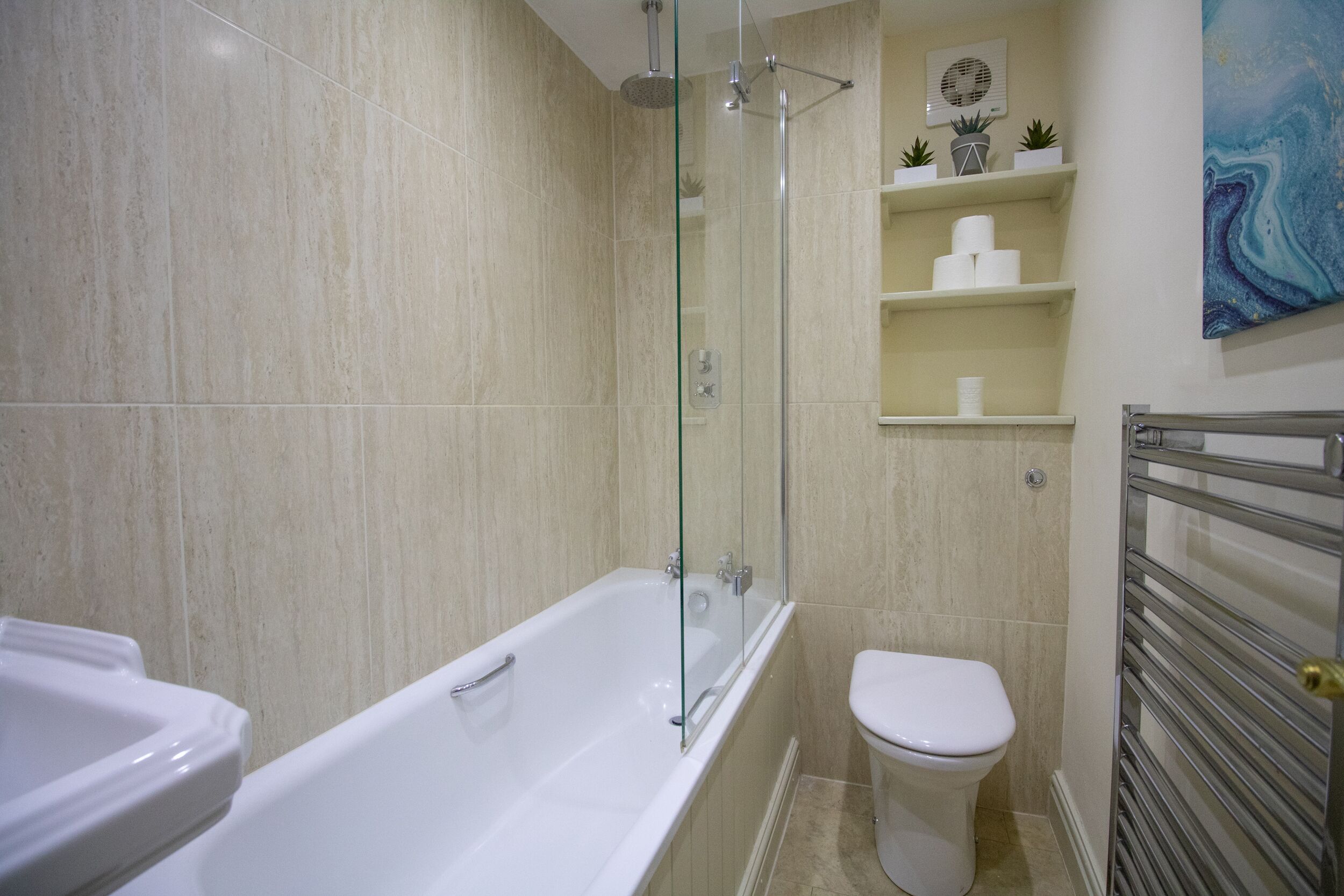 Et badeværelse med badekar, toilet og brusekabine med glasvægge. Der er hylder med toiletartikler synlige.