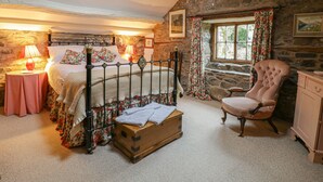 Ein rustikales Schlafzimmer mit einer Steinwand, einem hölzernen Bett mit floraler Bettwäsche, einem alten Stuhl und einer kleinen Holzablage.