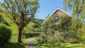 Ein Haus mit einem steinernen Weg, der zu einer hölzernen Bank führt, umgeben von üppiger Vegetation und einer hügeligen Landschaft.