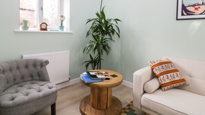 Un salon avec un fauteuil gris, un canapé blanc, une table basse en bois, une grande plante verte et une fenêtre donnant sur un immeuble en briques.