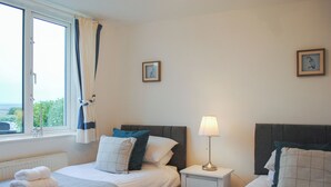 A hotel room with two beds, a window with curtains, a bedside table with a lamp, and a radiator. - Fairview (Salcombe)