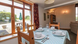 Un coin repas avec une table dressée pour quatre personnes, des chaises en bois, une nappe bleue à pois et une vue sur l’extérieur grâce à de grandes fenêtres.