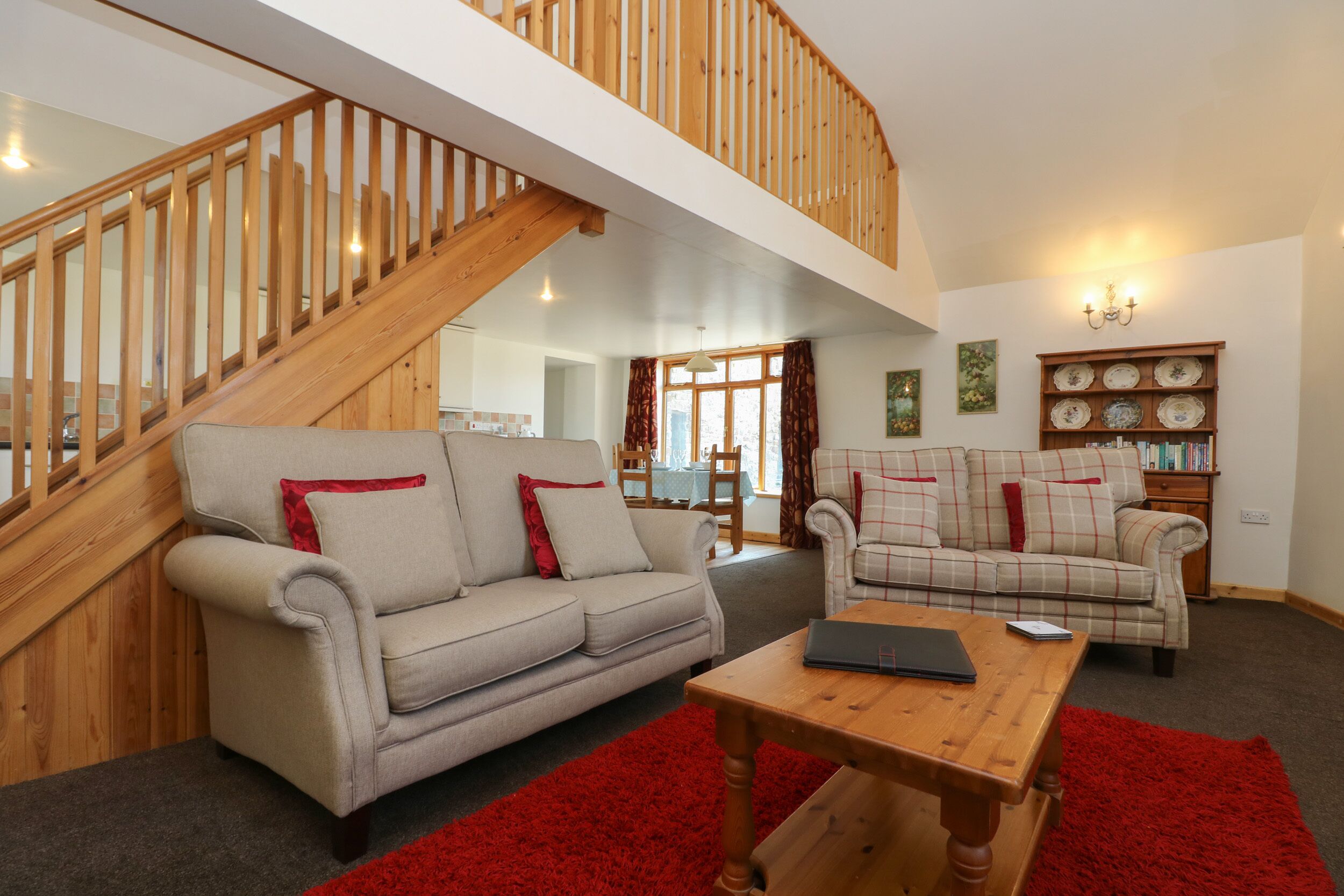 A spacious living room with a high wooden staircase, two sofas with red cushions, a wooden coffee table, and a bookshelf with decorative plates.