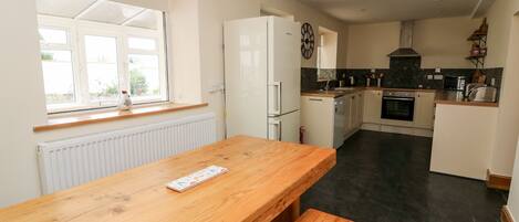 A compact kitchen with a wooden table and bench, a white refrigerator, a clock, and a window with a view.