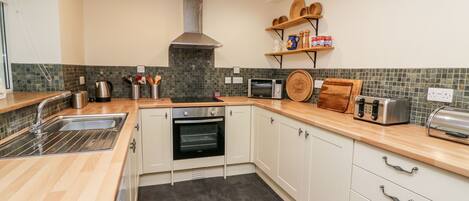 A modern kitchen with a stainless steel sink, a built-in oven, a microwave, and a toaster. There are wooden shelves above the counter holding dishes and a bread box.
