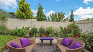 Un patio ajardinado con mobiliario de mimbre y una mesa, rodeado de diversas plantas y flores.