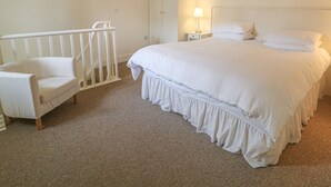 A bedroom with a large bed, a white chair, a nightstand with a lamp, and a wardrobe.