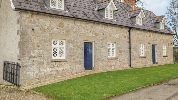 A stone building with a row of windows and doors, a grassy lawn, and a paved driveway.
