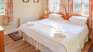 Una cama bien hecha con sábanas blancas y una toalla doblada, una mesita de noche de madera con una lámpara, y dos cuadros en la pared.