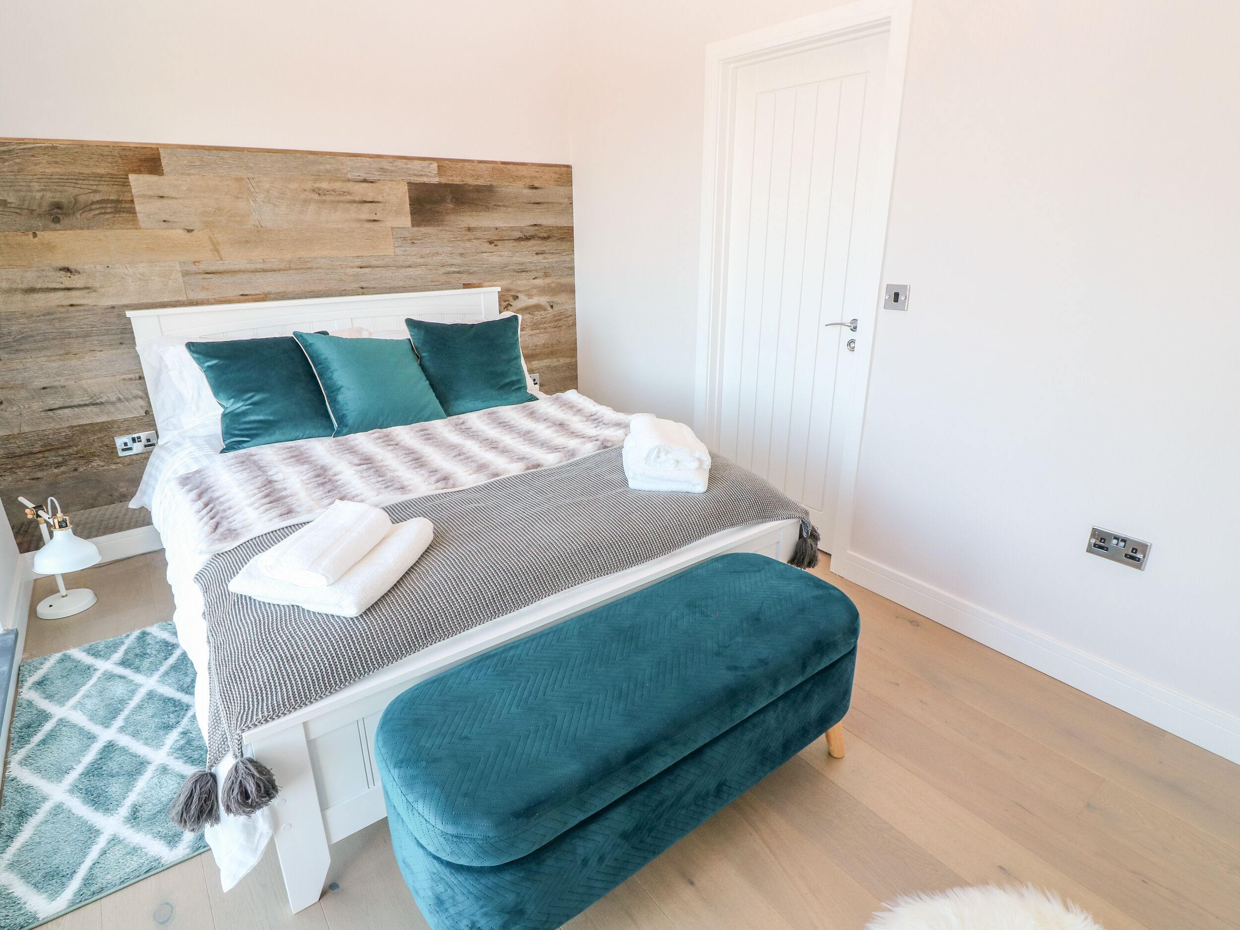 A neatly arranged bedroom with a wooden headboard, a bed with teal pillows, a grey and white striped blanket, and a teal bench.