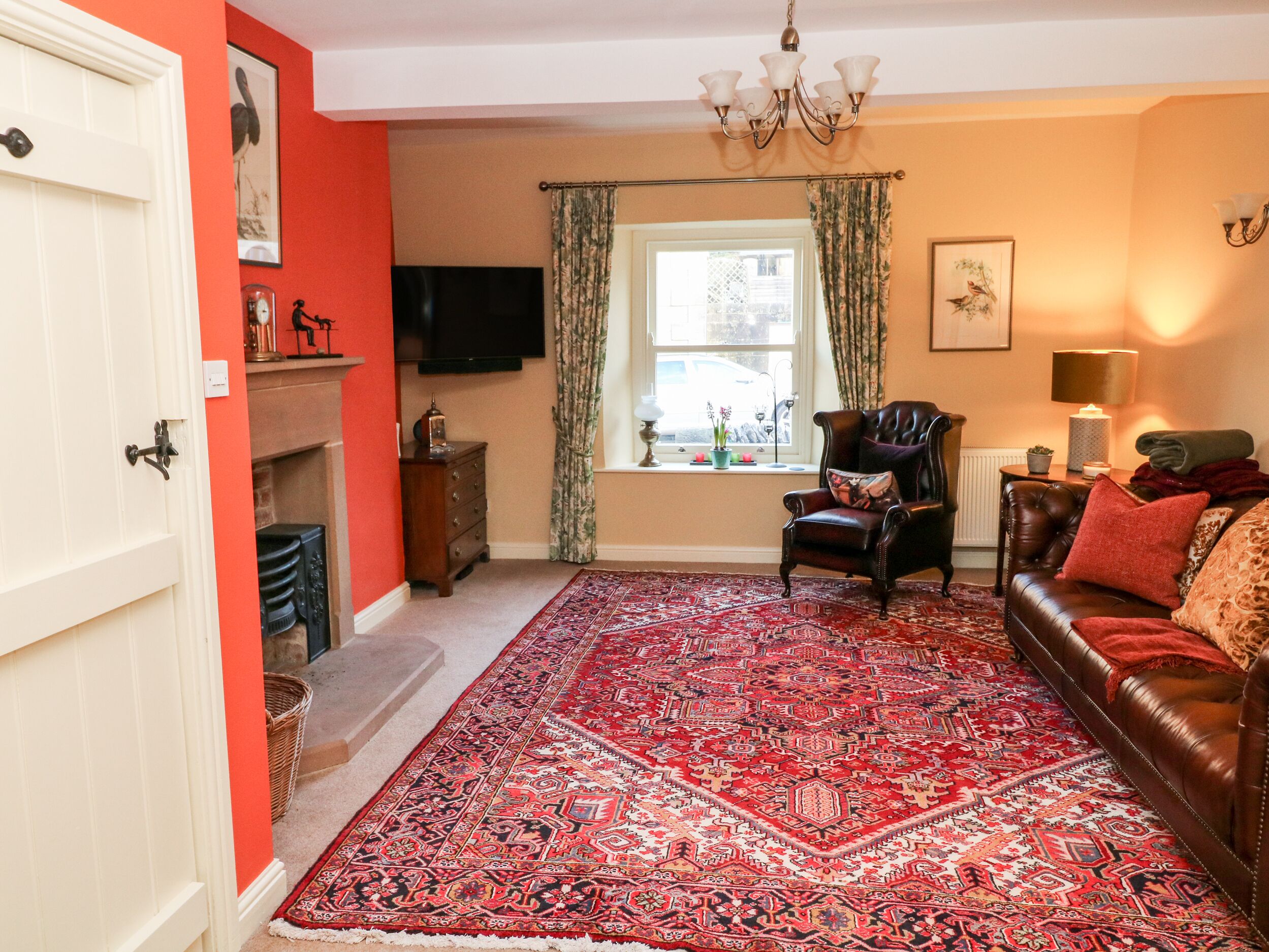 Un salón con alfombra roja, chimenea, televisor, ventana con cortinas, sofá y candelabro.