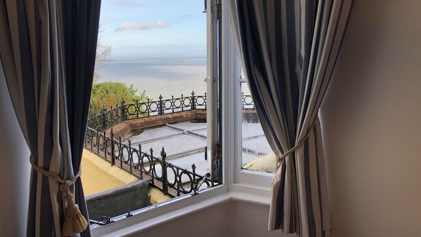 A window with striped curtains, a view of a coastal area with a fence and greenery. - Blue Bay (Lynton)