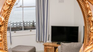 A room with a TV on a wooden stand, a grey sofa, a small ottoman, a window with a view of the sea, and a striped curtain. - Blue Bay (Lynton)