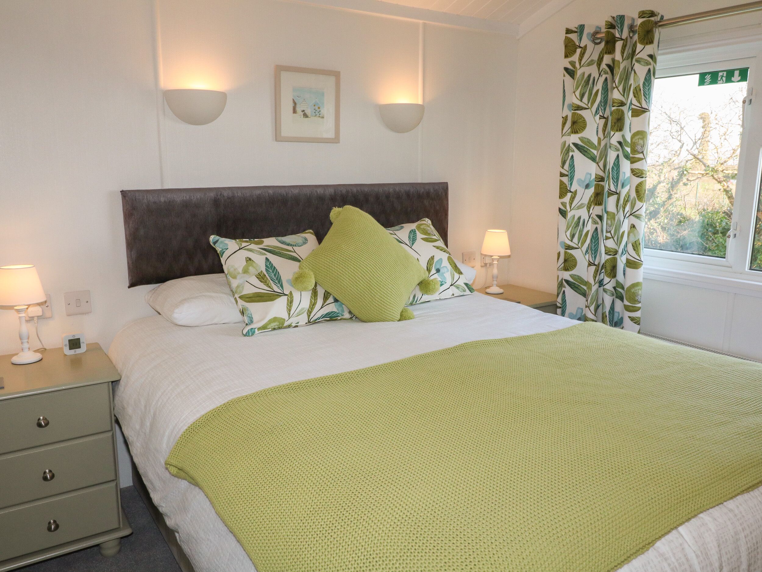 A neatly made bed with a green throw blanket, two bedside lamps, a nightstand with a digital clock, and a window with a view of trees.