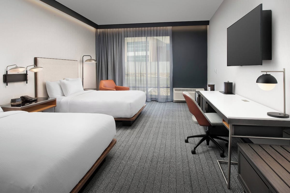 Ein Hotelzimmer mit zwei Betten, einem Schreibtisch, einem Stuhl, einem Fernseher und einem großen Fenster mit Vorhängen.