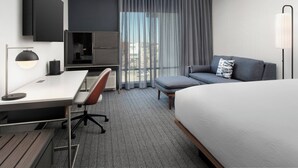 Una habitación de hotel moderna con cama, escritorio con silla, sofá, televisor y un ventanal con cortinas.