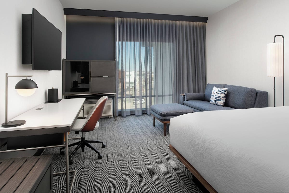 Ein modernes Hotelzimmer mit einem Bett, einem Schreibtisch mit Stuhl, einer Couch, einem Fernseher und einem großen Fenster mit Vorhängen.